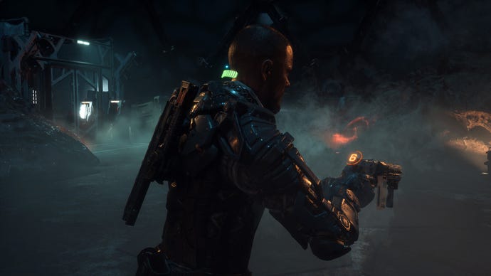 The Callisto Protocol's main character, wearing futuristic armor and holding a weapon, walking through a dank dimly lit area.